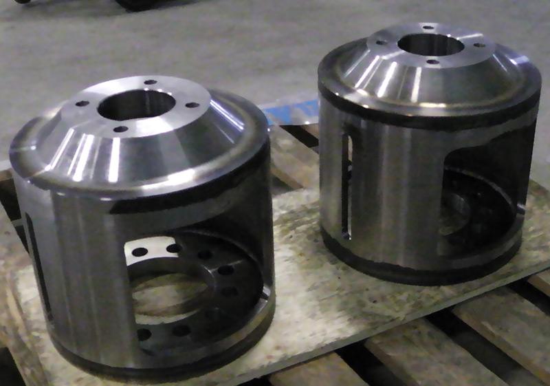 Two cylindrical, machined steel industrial parts with large side cutouts, sitting on a wooden pallet in a workshop.