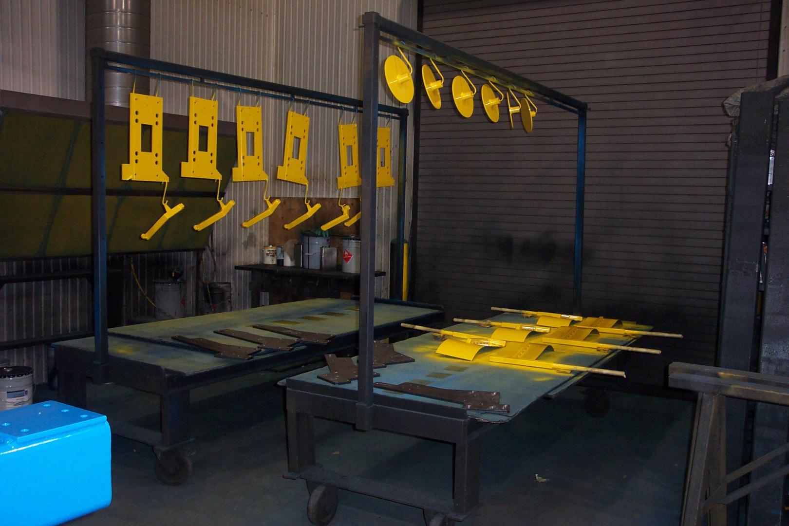 Yellow metal parts hang from a rack and lie on tables in an industrial workshop.