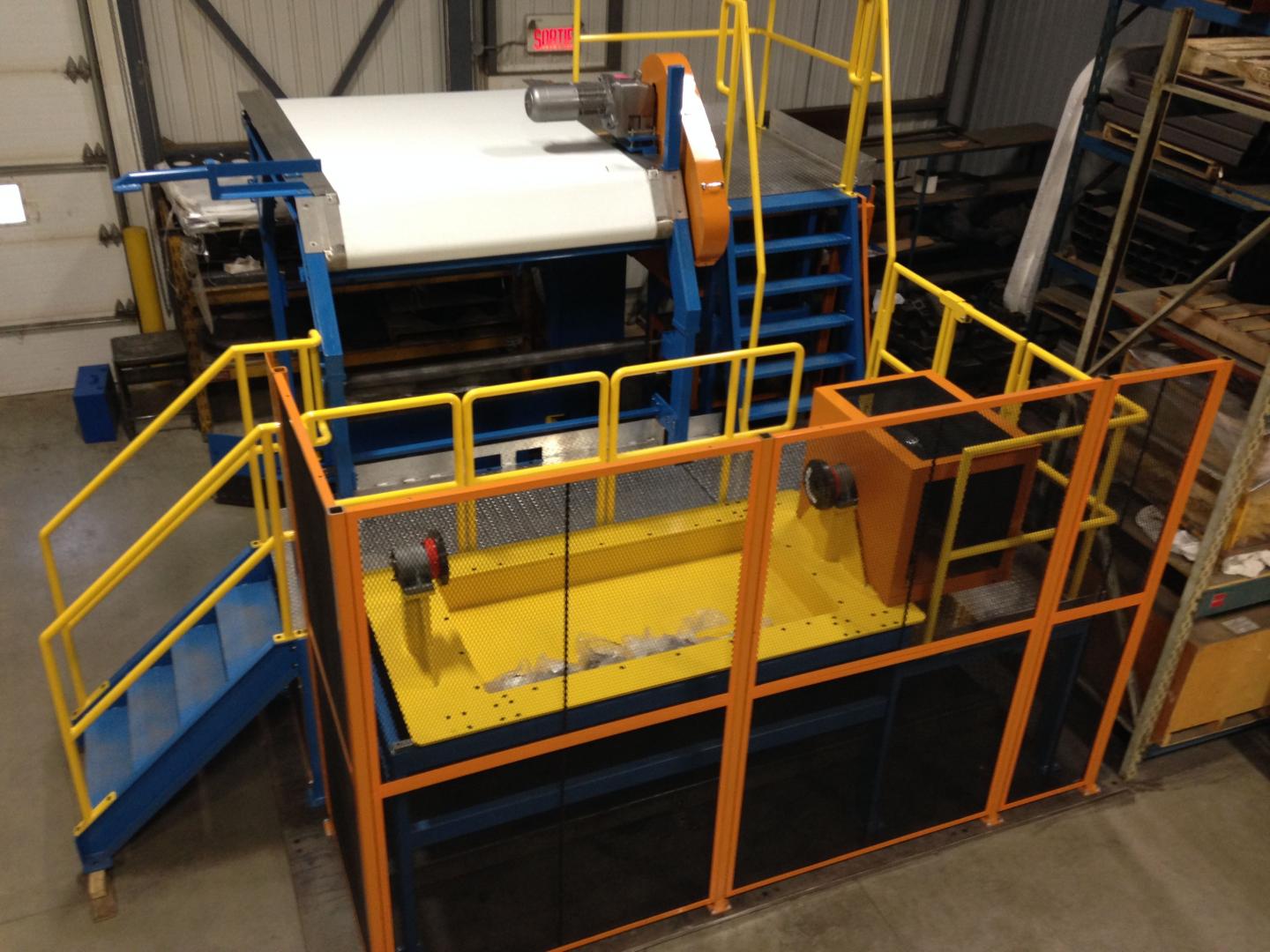 An industrial machine with blue framework, yellow safety railings, and a white sheet being processed in a facility.