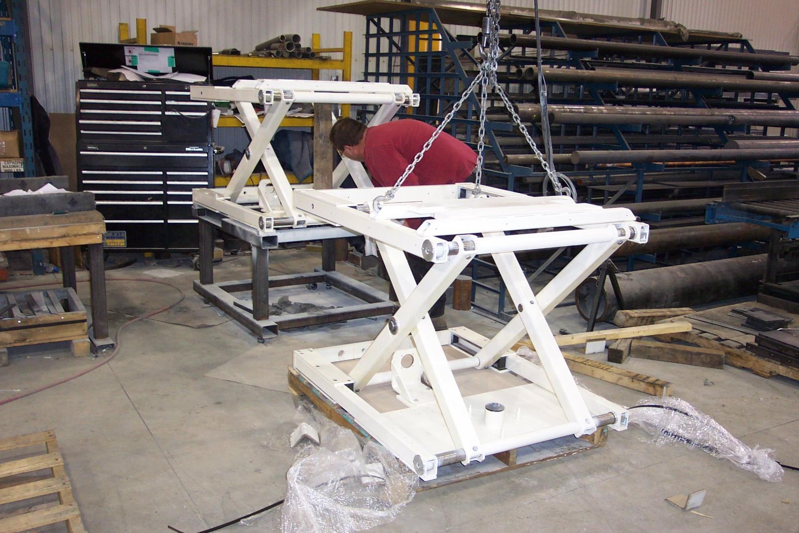 A person in a red shirt works in a workshop, positioning a large white industrial scissor lift frame with a crane.