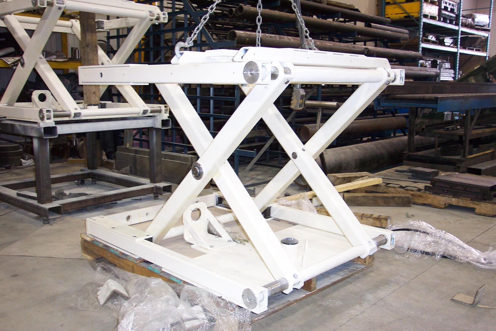 A white, metal scissor lift mechanism on a pallet inside a warehouse, with a second unit partially visible behind it.
