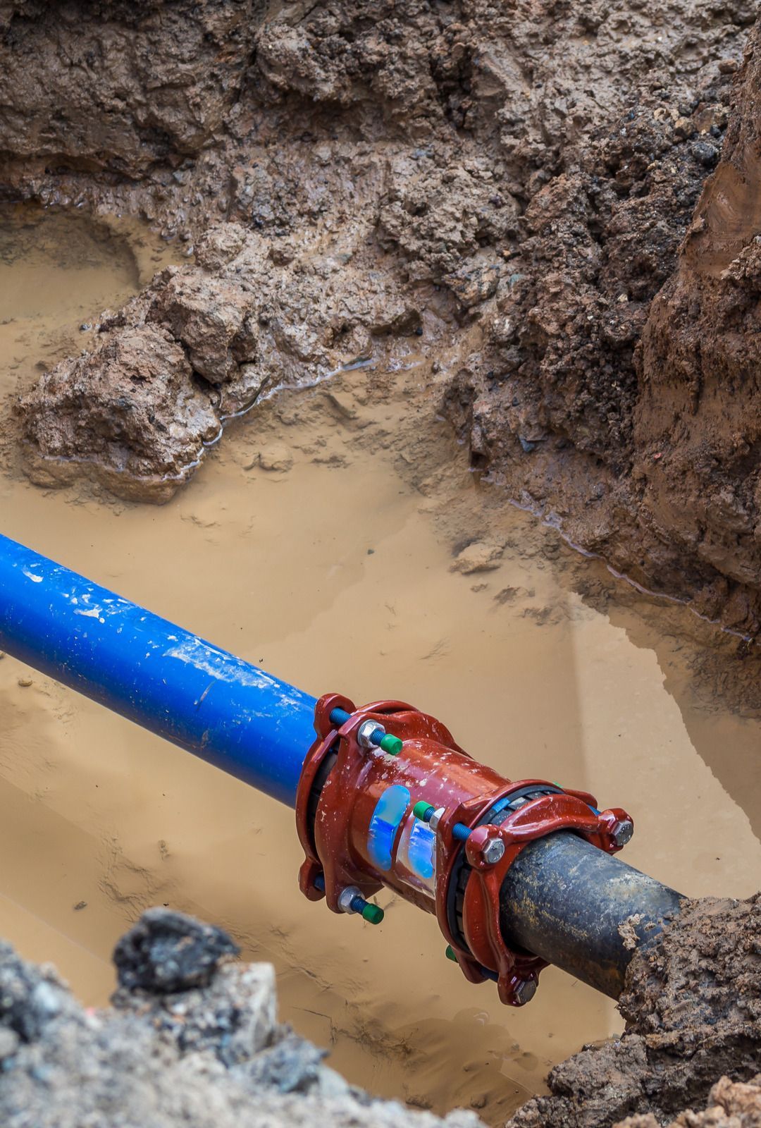 A blue pipe connected to a dark pipe via a large red coupling fitting within a muddy construction trench.