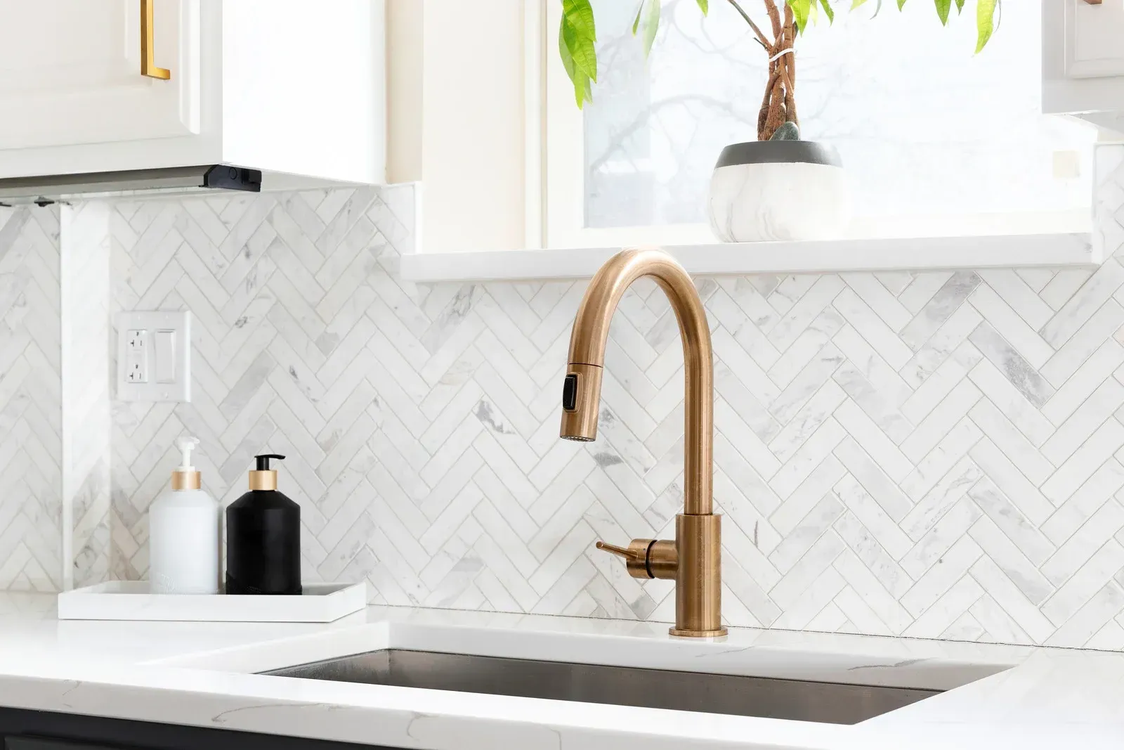 A gold-toned kitchen faucet stands above a white marble countertop against a white herringbone-tiled backsplash.