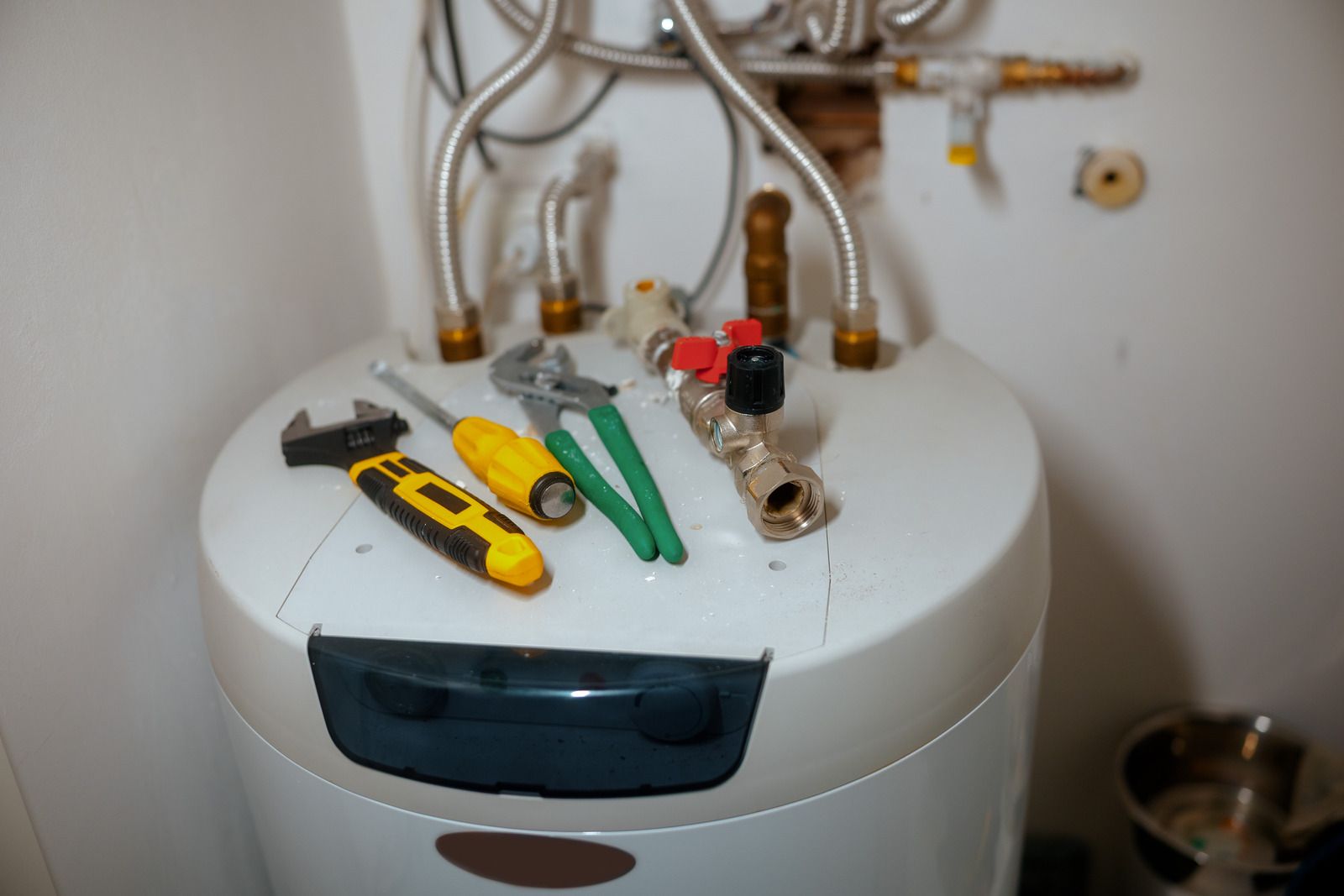 Tools including a wrench, screwdriver, and pliers rest on top of a water heater with exposed plumbing connections.