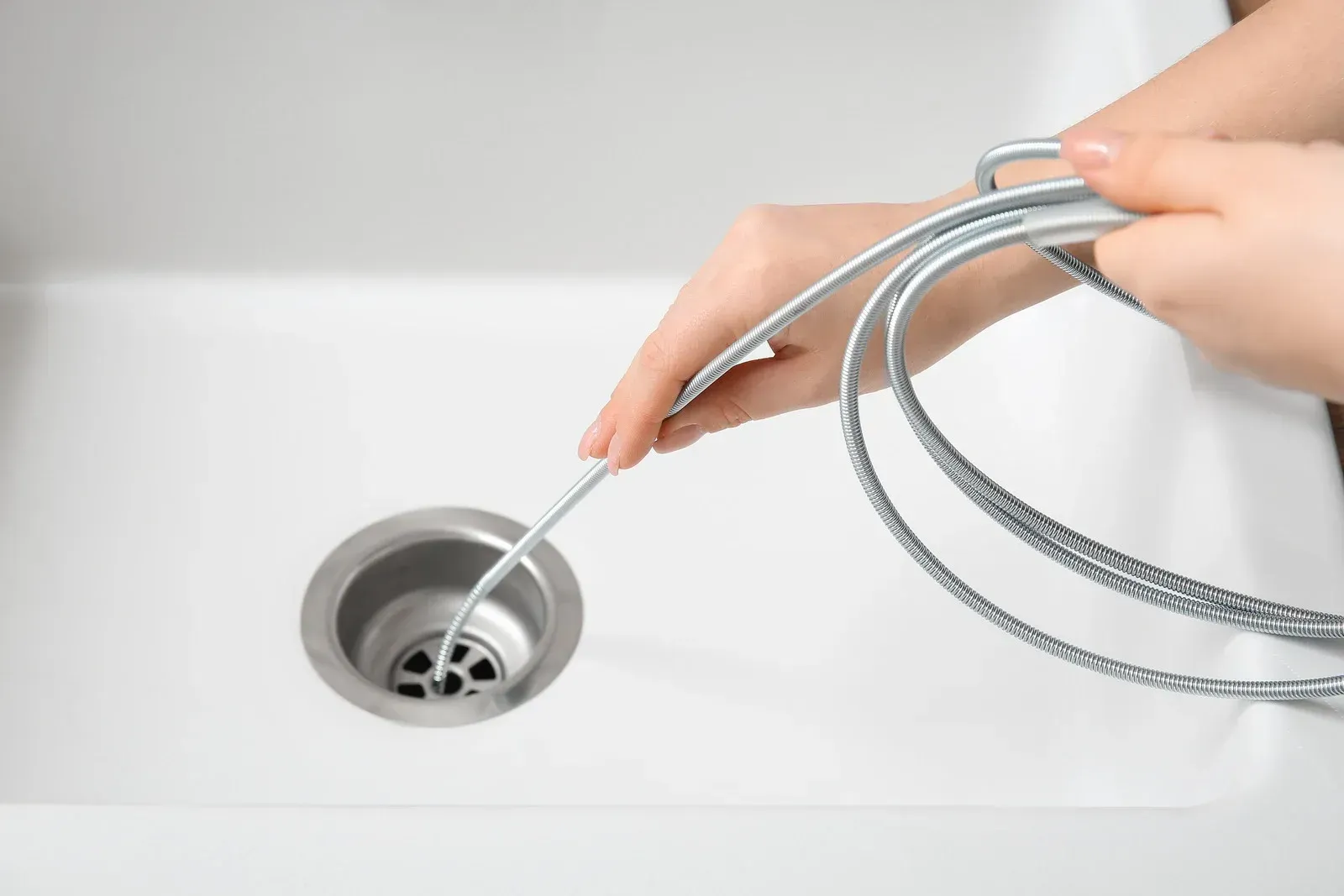 A person uses a thin, silver metal drain cleaning tool to clear a clog in a white kitchen sink.