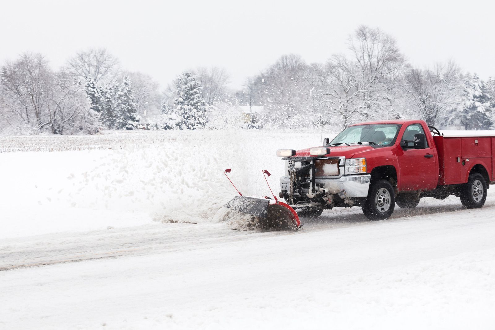 A red snowplow truck clearing a snowy road with a white, wintry field and trees in the background.