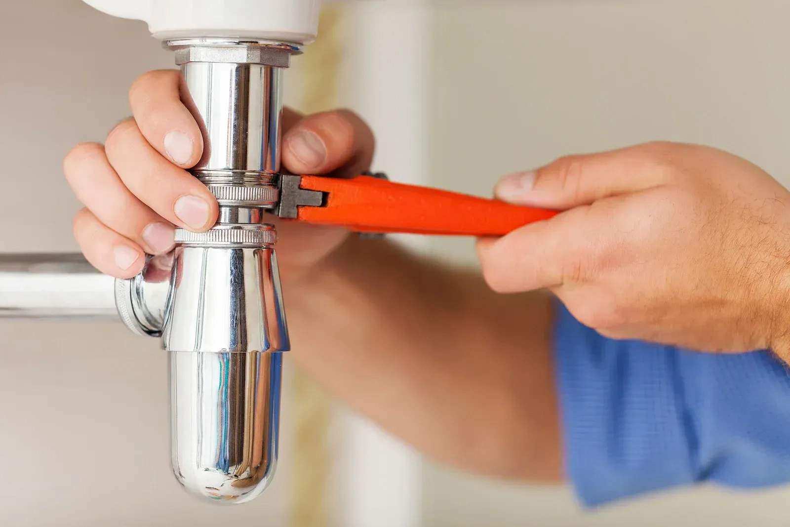 Hands using an orange wrench to tighten a chrome pipe fitting on a sink drain.
