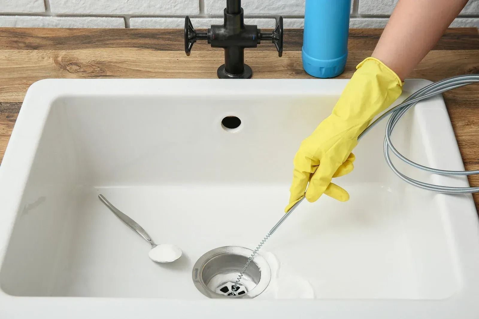 A person wearing a yellow rubber glove uses a drain snake to clean a white kitchen sink.