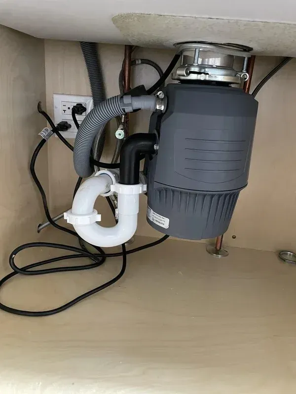 A grey garbage disposal unit installed under a kitchen sink with visible plumbing and a power cord plugged into an outlet.