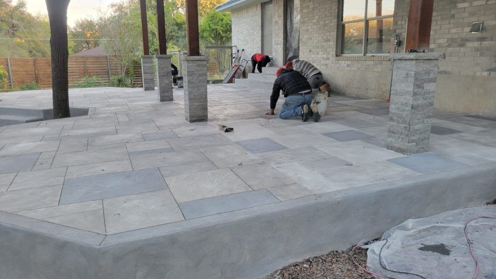 Two workers install pavers on a new gray stone patio featuring stone columns and steps in front of a house.