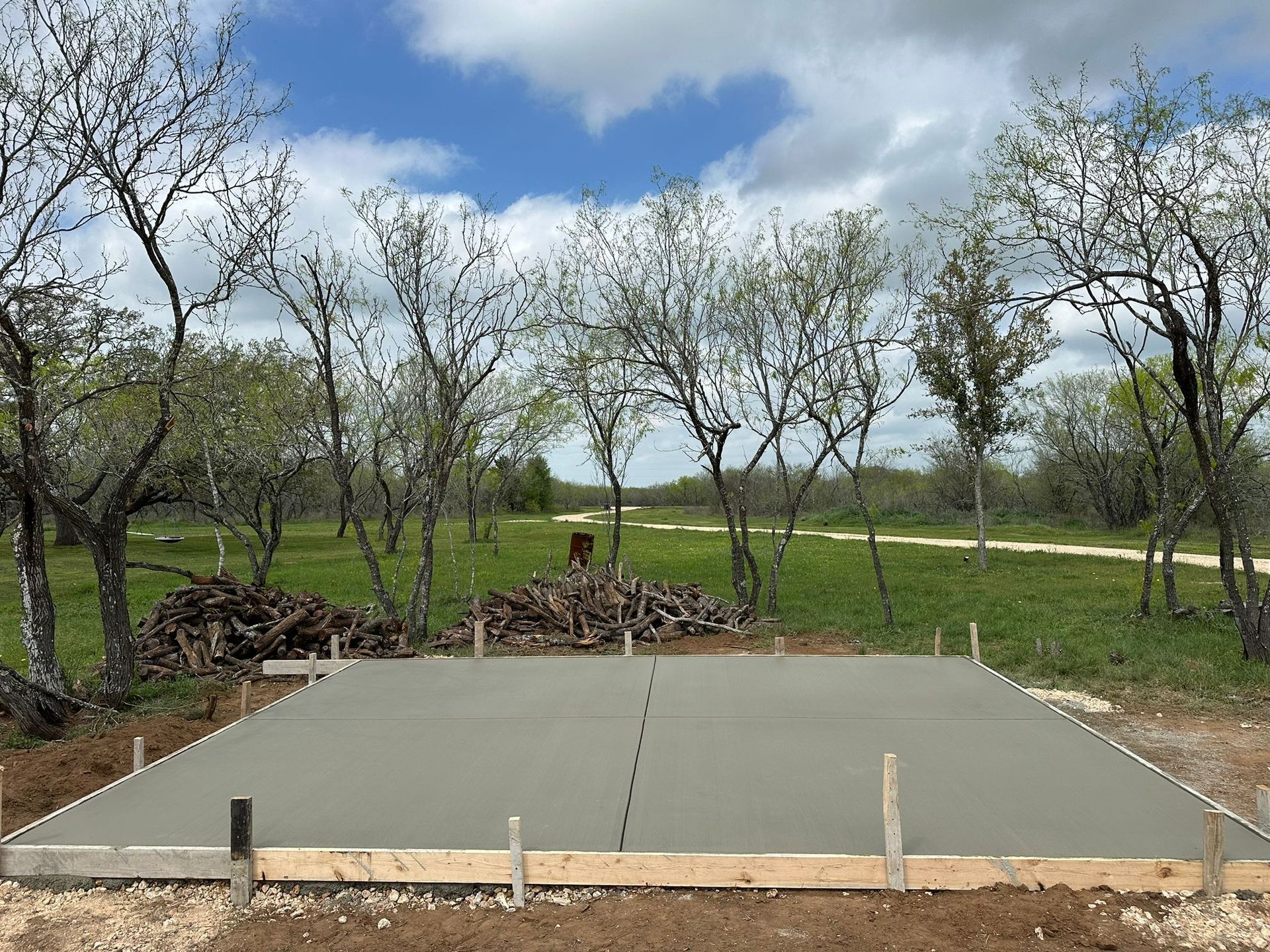 A fresh concrete slab foundation for a building project set in a grassy clearing with trees and a dirt path behind it.