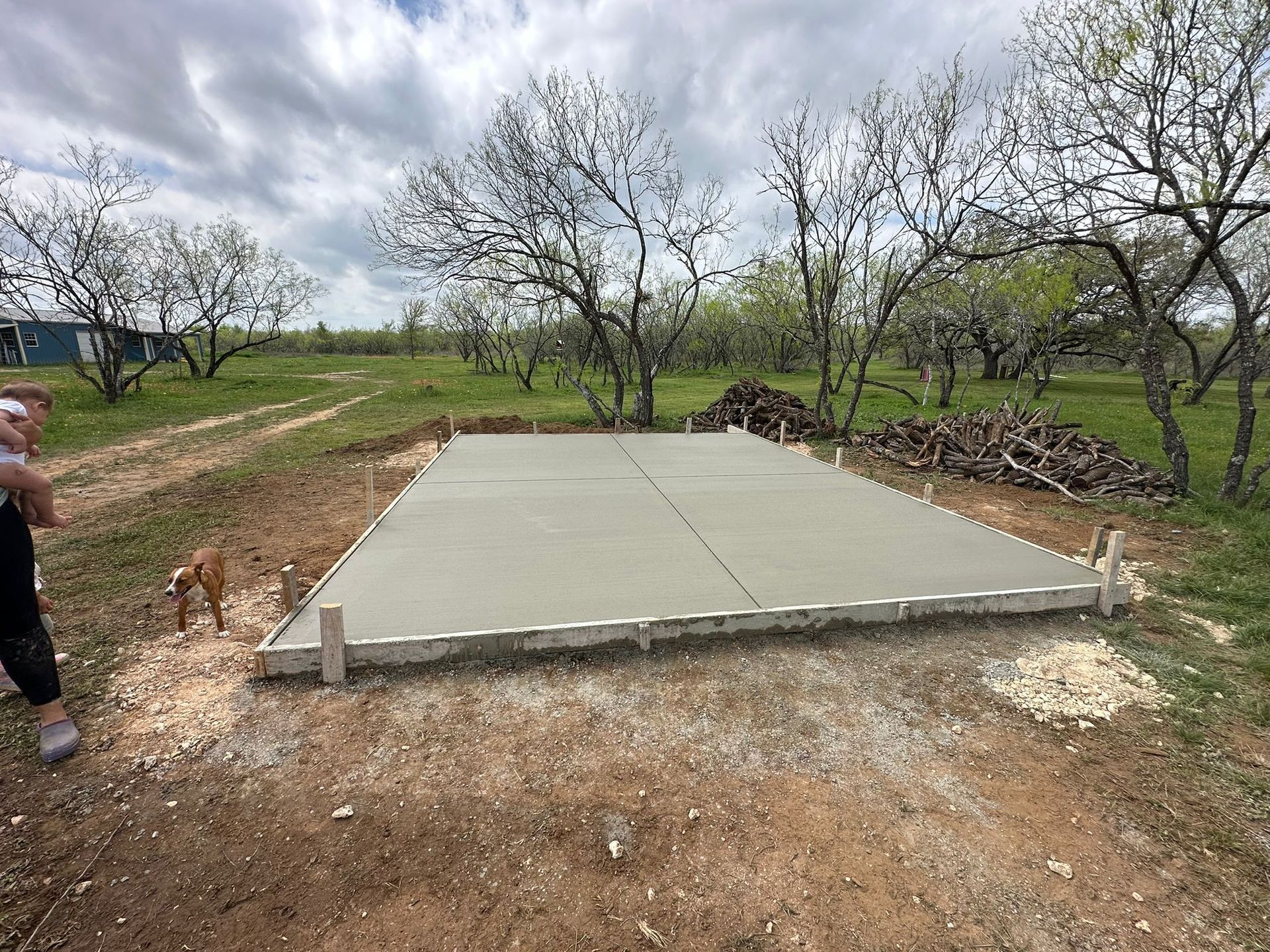 A freshly poured concrete slab sits in a rural outdoor setting with trees in the background and a dog nearby.