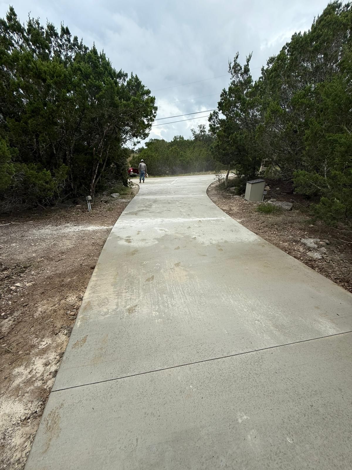 A concrete pathway winds through trees and shrubs with a person walking in the distance under a cloudy sky.