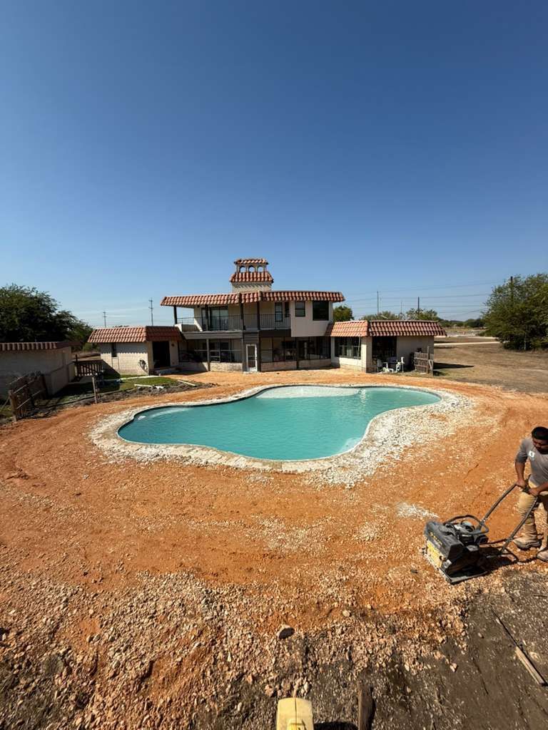 A person operates a compactor on a dirt lot surrounding a newly installed turquoise swimming pool in front of a house.
