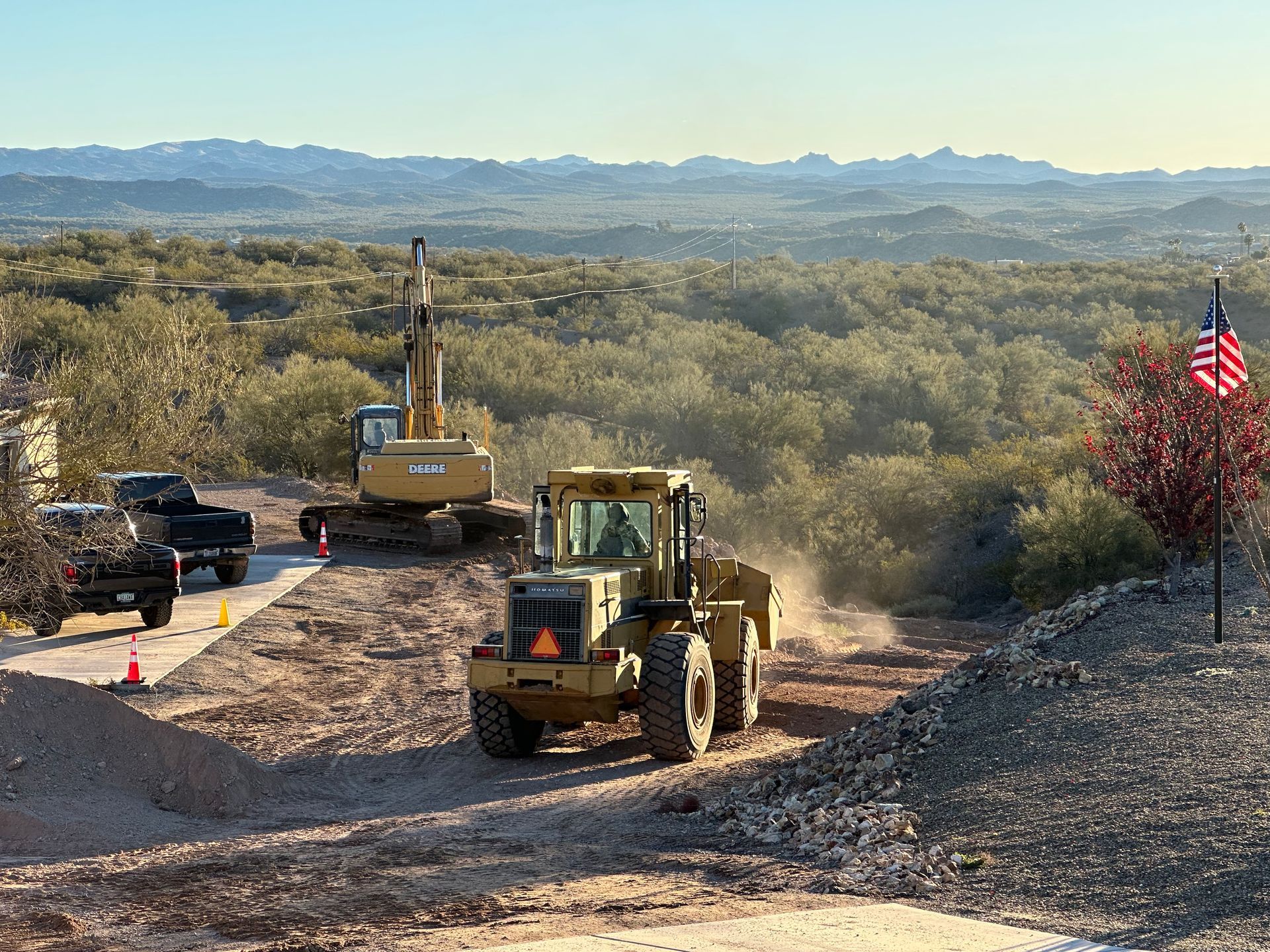 Roadwork with two construction vehicles on a dirt path in a desert landscape, U.S. flag at right.