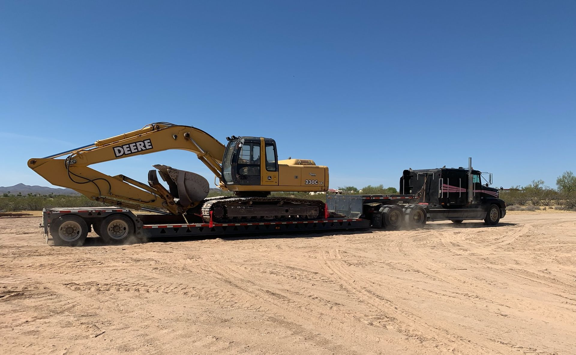 Yellow excavator on a flatbed trailer in a sandy construction site under a clear blue sky.