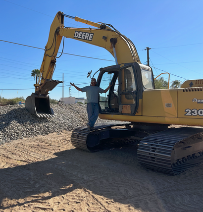 Worker guiding a John Deere excavator on a gravel site under a clear blue sky