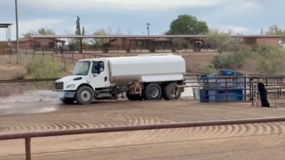 White water truck spraying a dirt track beside a blue metal equipment trailer in a dry outdoor area