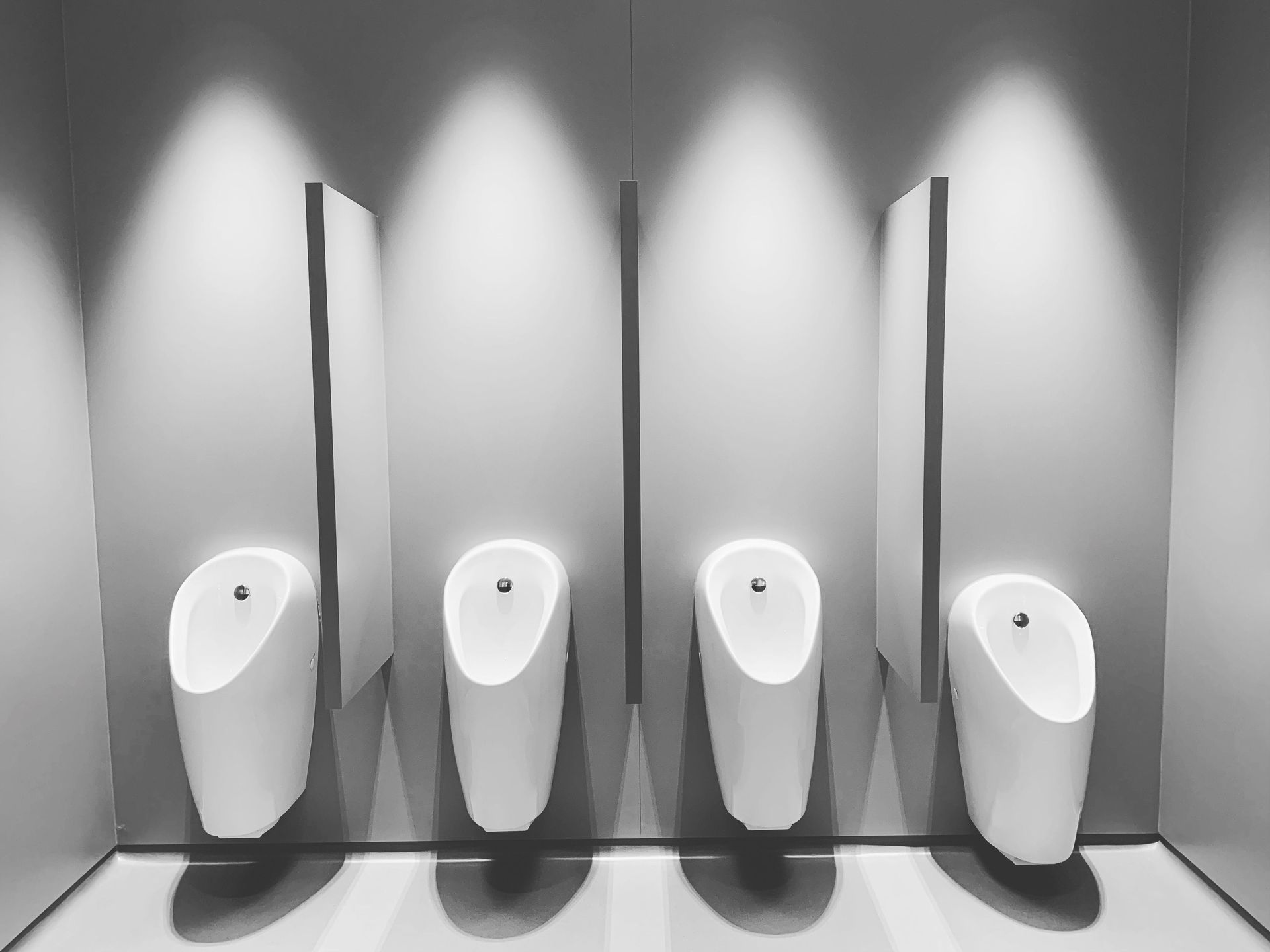Four urinals along a gray wall, separated by a thin divider and illuminated by modern lighting.