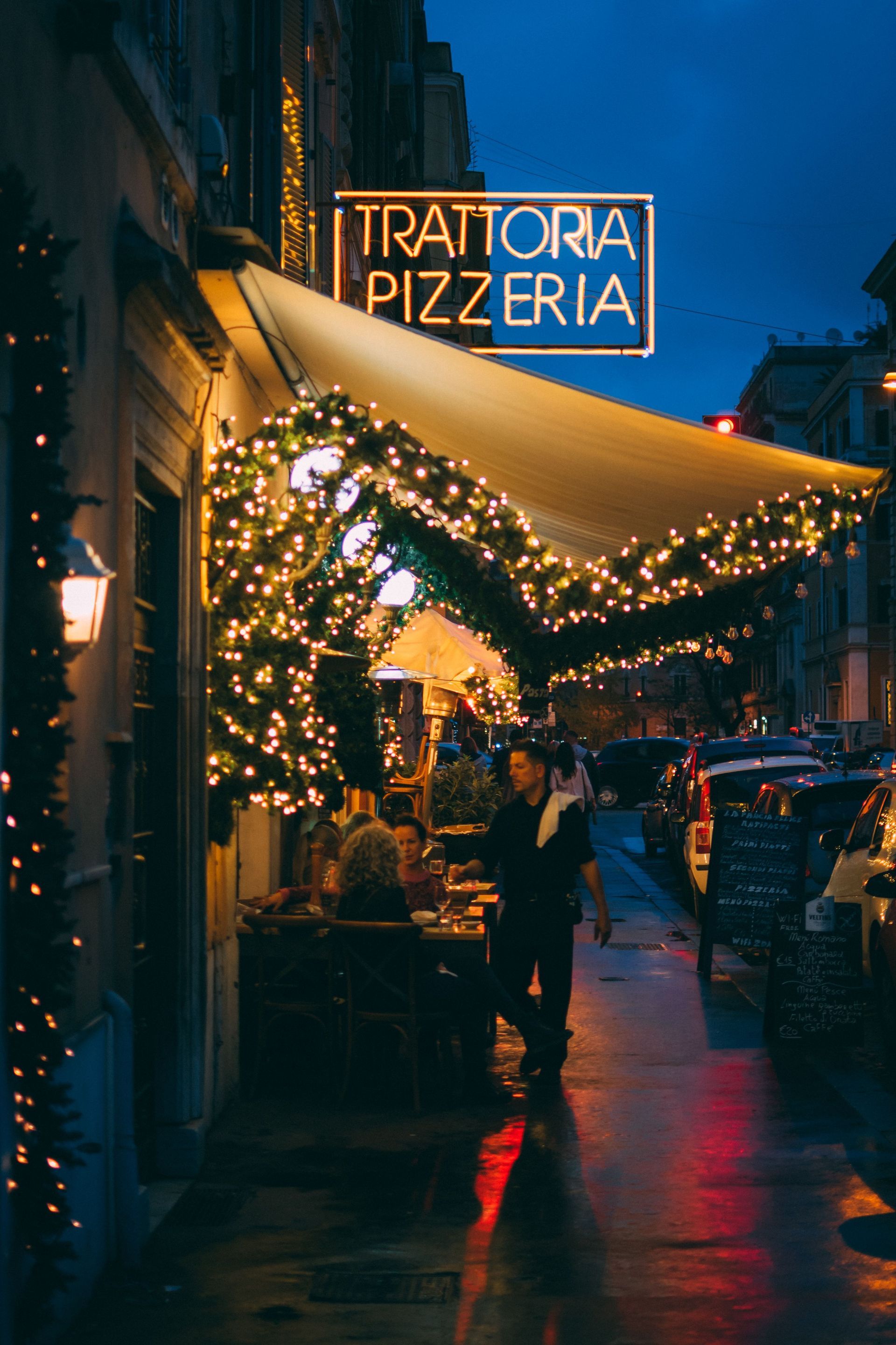 Side view of the entrance of a pizzeria with guests dining outdoors and a waiter attending to them.
