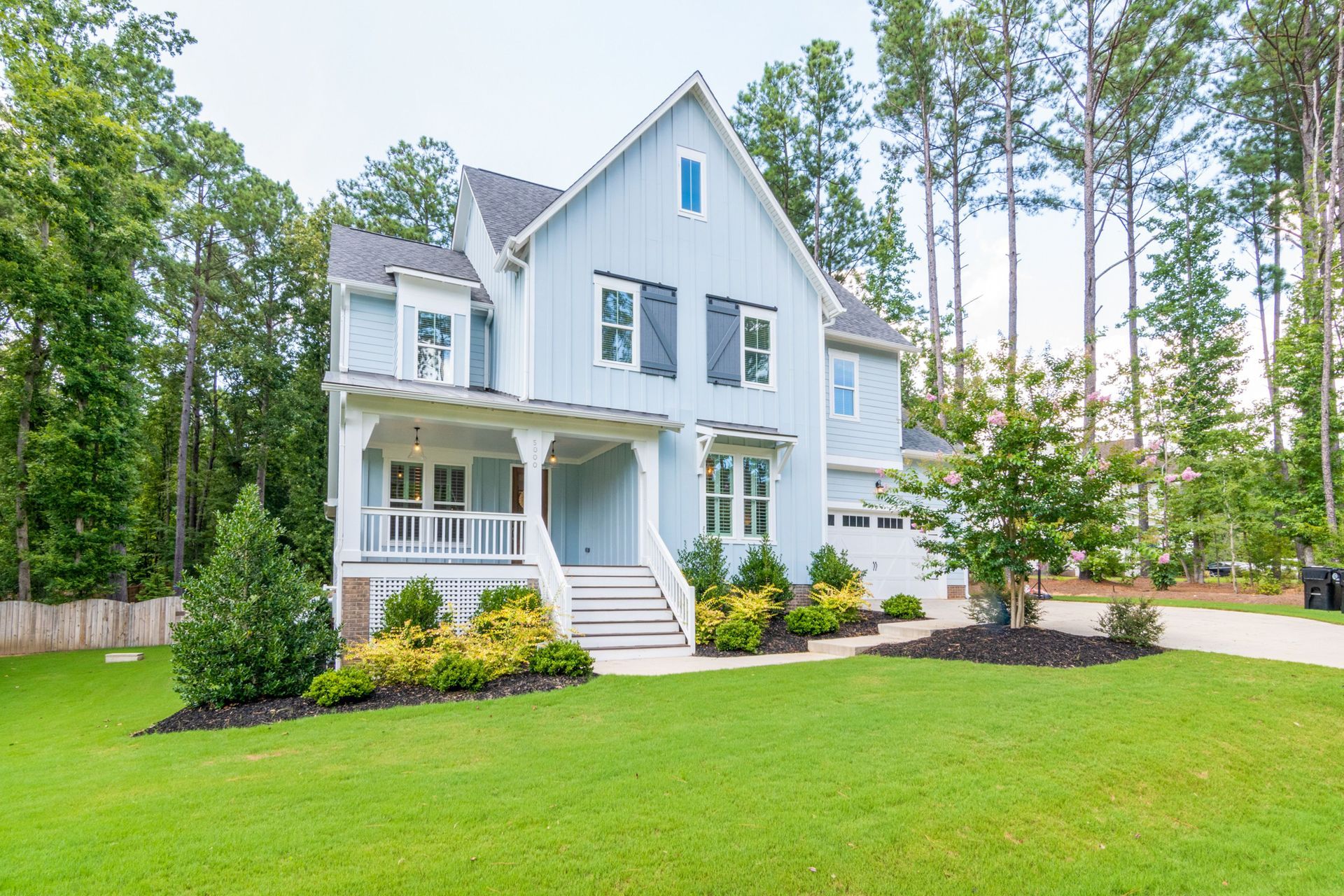 Two-story, modern, suburban single family home with large front yard, gardening, and a tree with forested area behind the house.