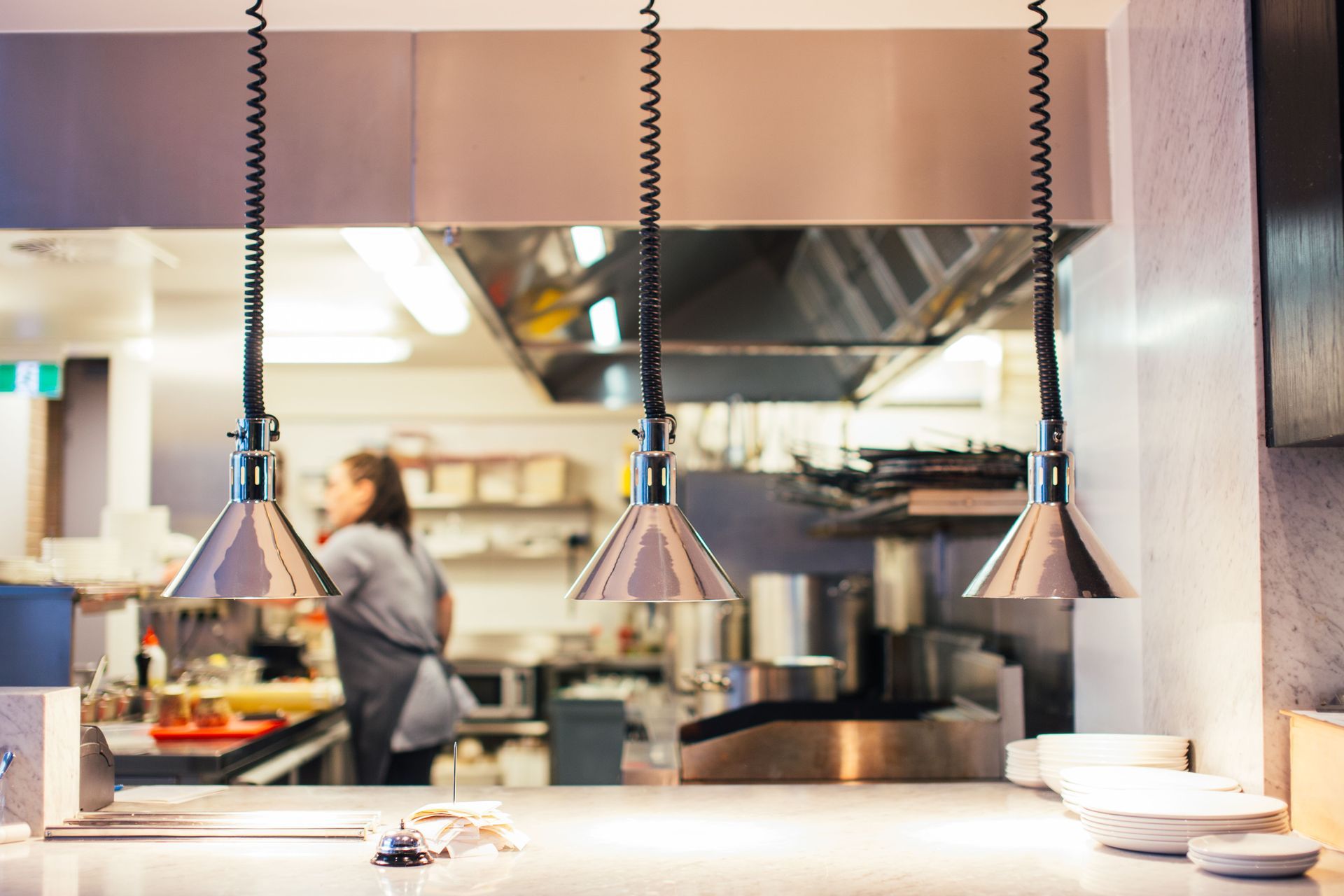 Commercial kitchen with employee in the background and a bell and customer tickets on the front counter.
