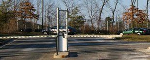 Parking lot entrance gate with a keypad post, trees, and parked cars in the background