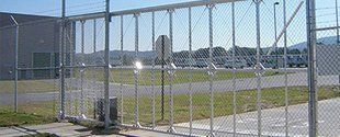 Chain-link fence in front of an open grassy field with a paved path and industrial buildings in the background