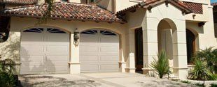 Spanish-style house with two beige garage doors, tiled roof, arched entry, and a sunlit driveway