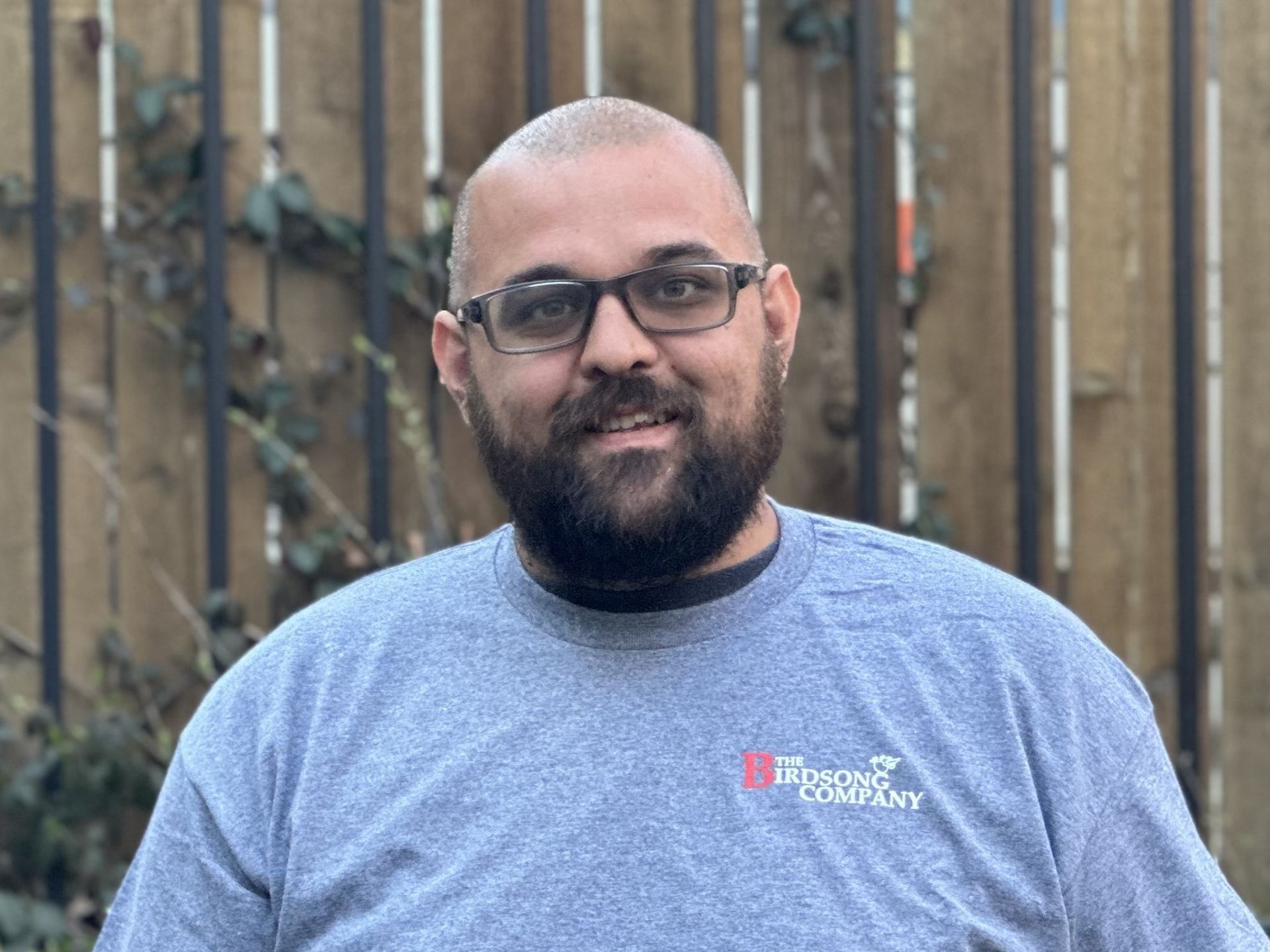 Smiling bald man with glasses in a blue T-shirt standing outdoors against a wooden fence