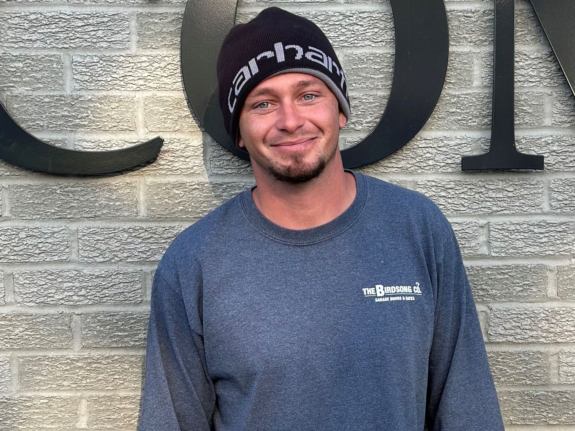 Smiling man in a black beanie and blue shirt standing against a gray brick wall with large letters behind him
