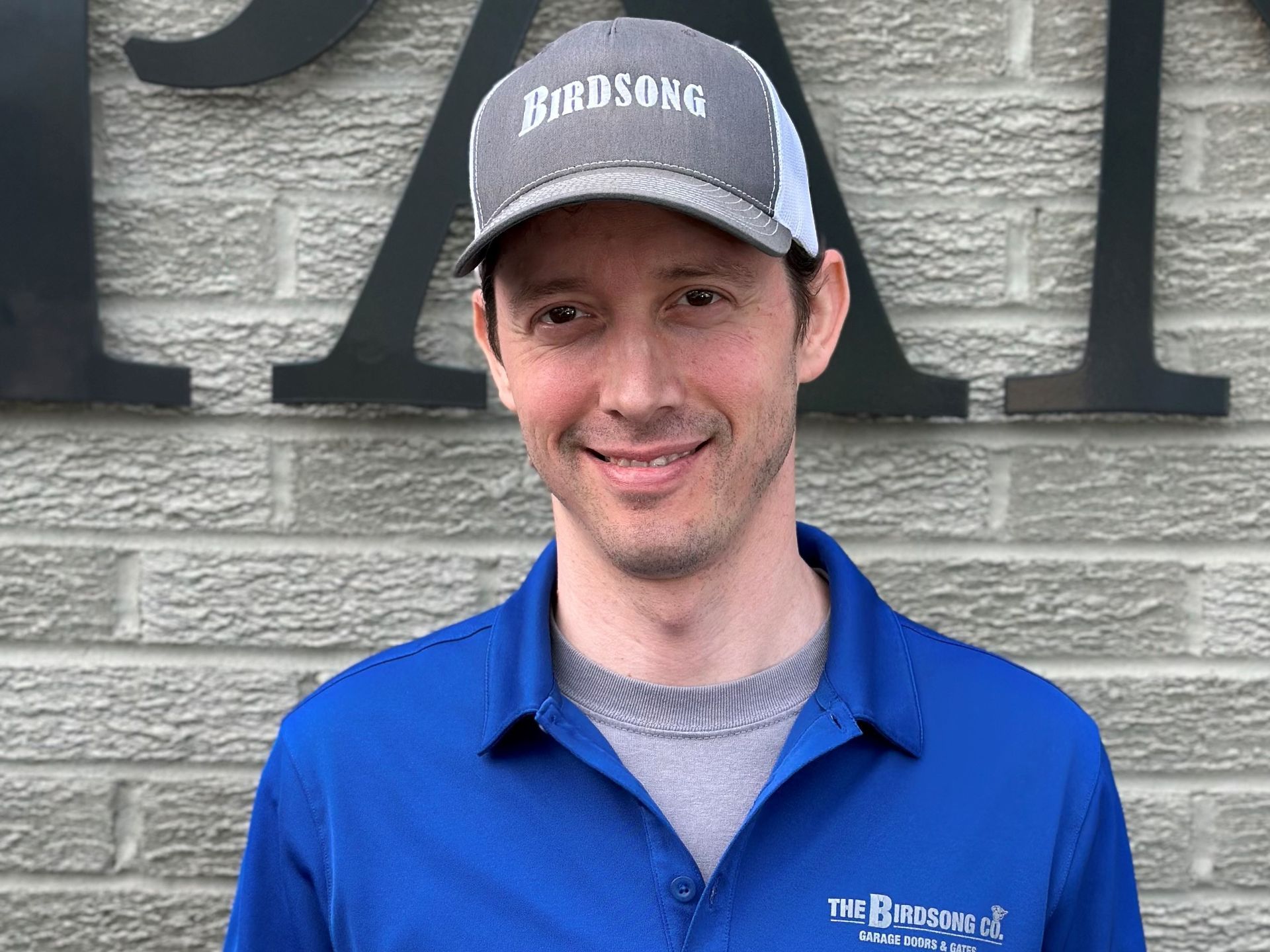 Smiling person in a blue shirt and gray cap standing in front of a brick wall