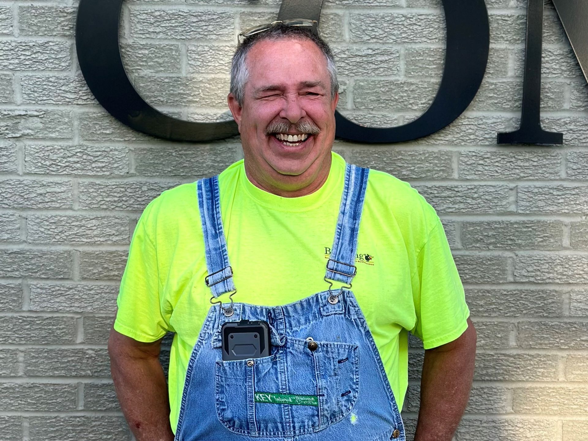 Smiling man in neon yellow shirt and blue overalls standing in front of a stone wall with large letters