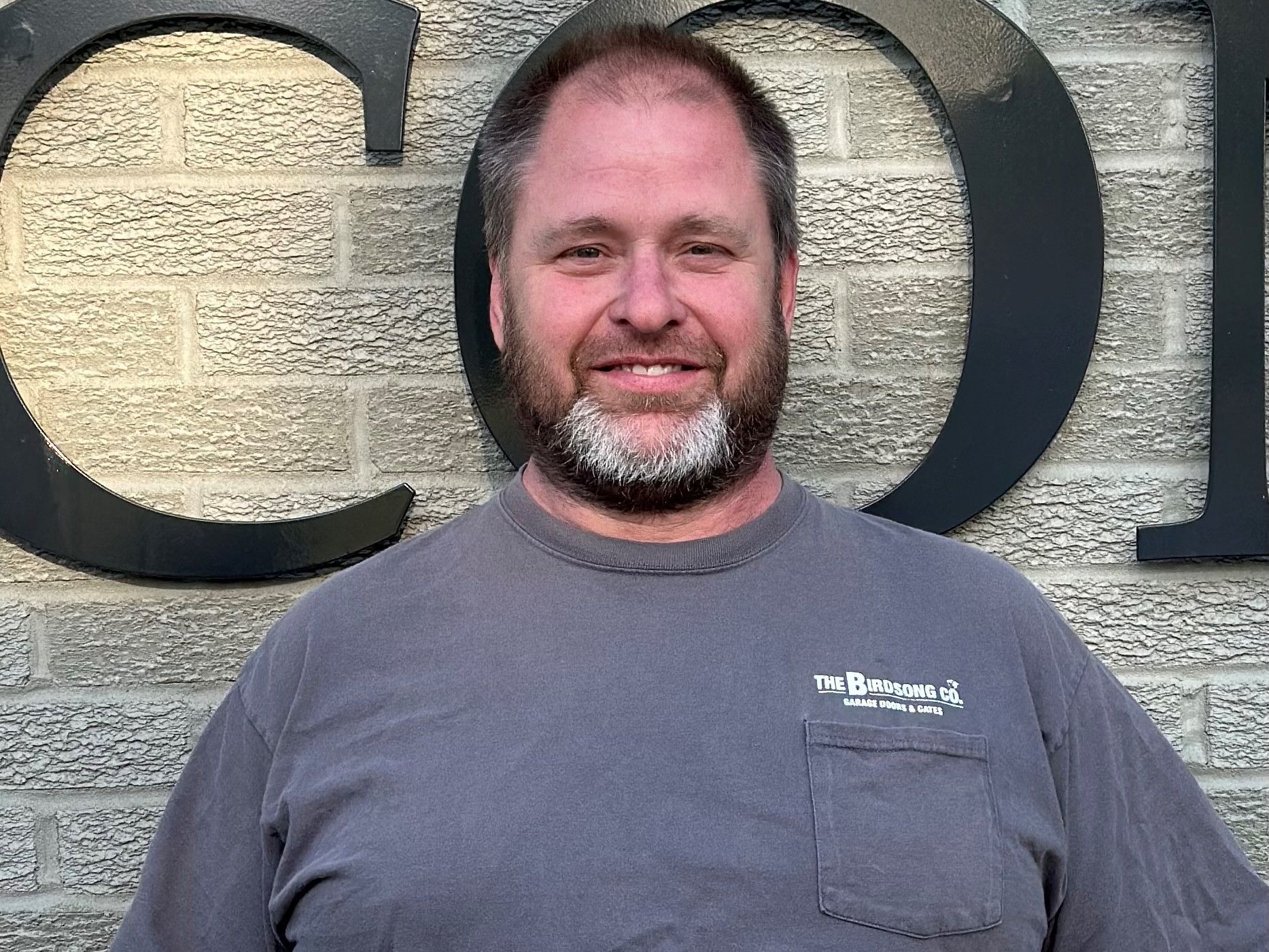 Man smiling in a gray shirt standing in front of a brick wall with large black letters