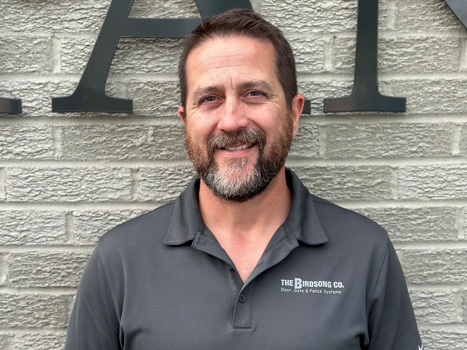 Smiling man in dark gray polo standing before a stone wall with large letters behind him