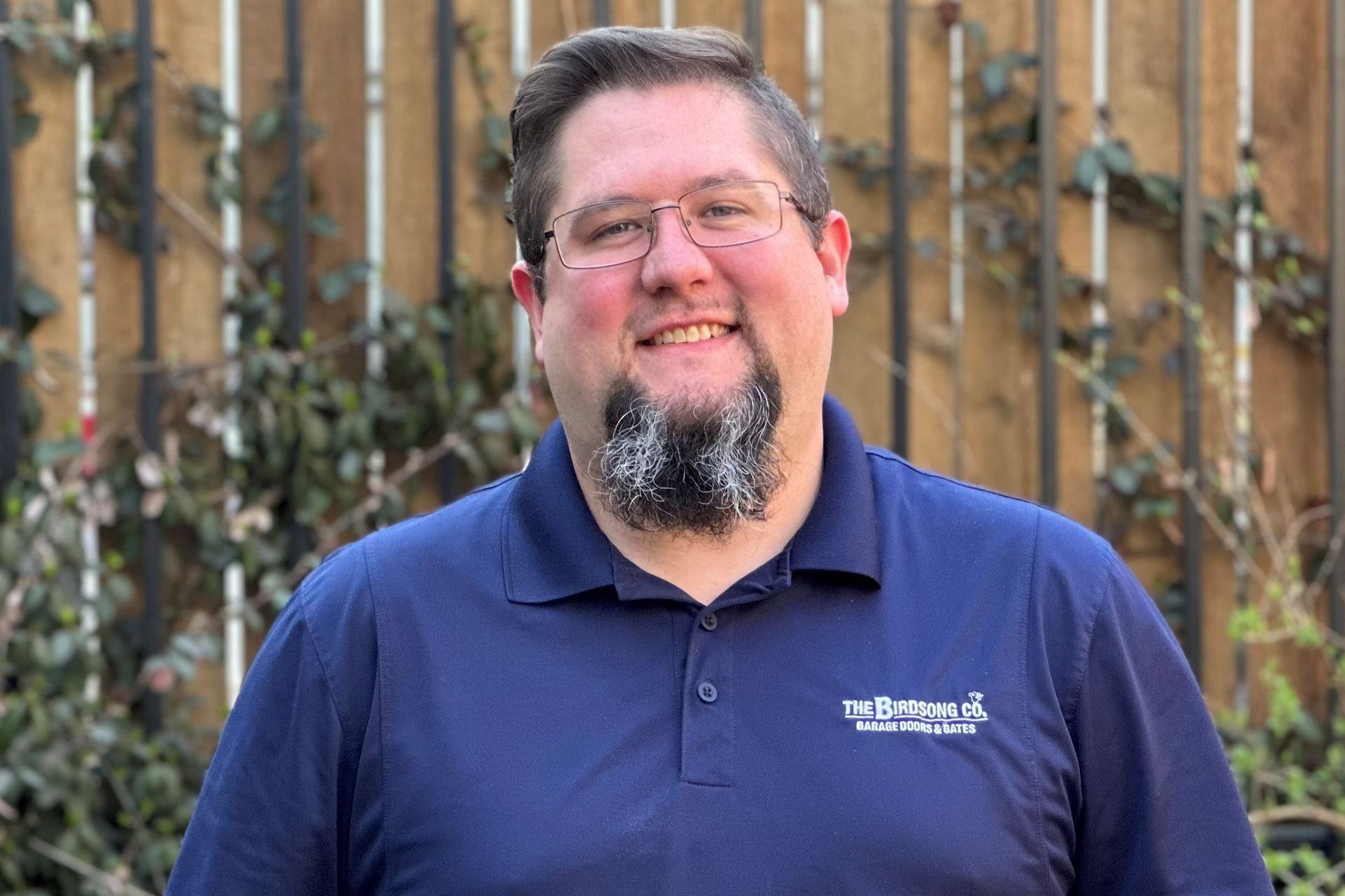 Smiling man in a navy polo shirt standing outdoors in front of a wooden fence and greenery