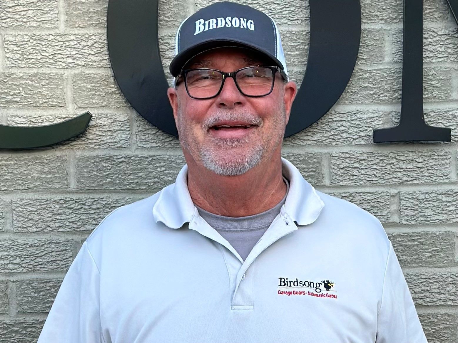 Smiling man in glasses and a Birdsong cap, wearing a white polo in front of a brick wall