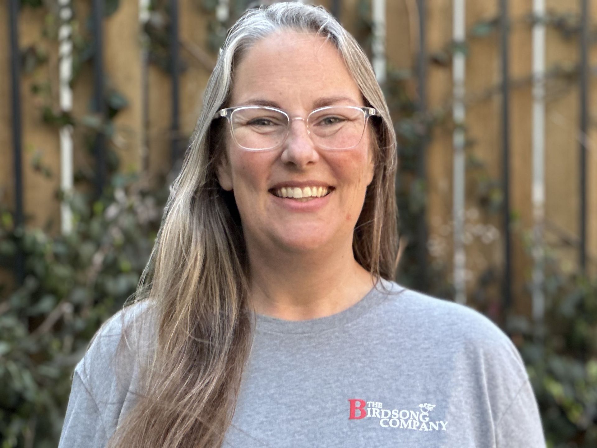 Smiling person with long gray hair and glasses wearing a gray T-shirt outdoors near a fence