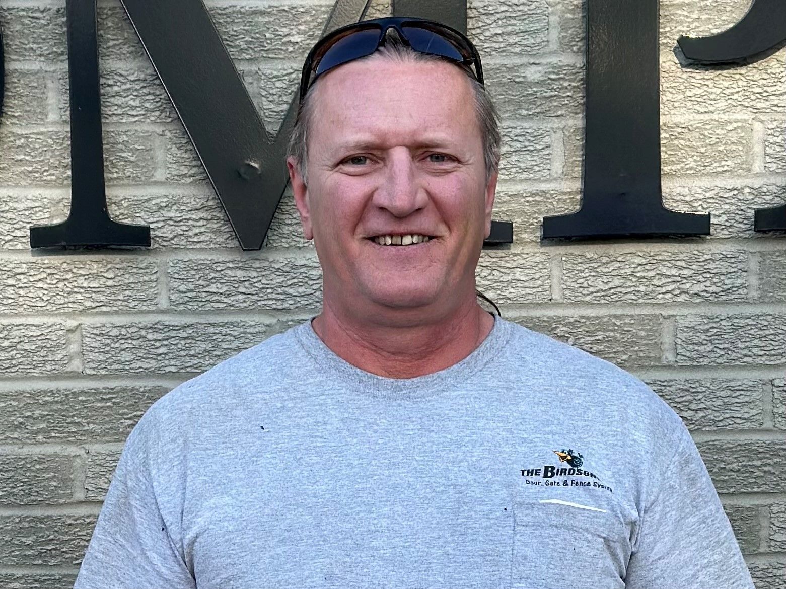 Smiling man in gray T-shirt standing against a stone wall with large black letters behind him