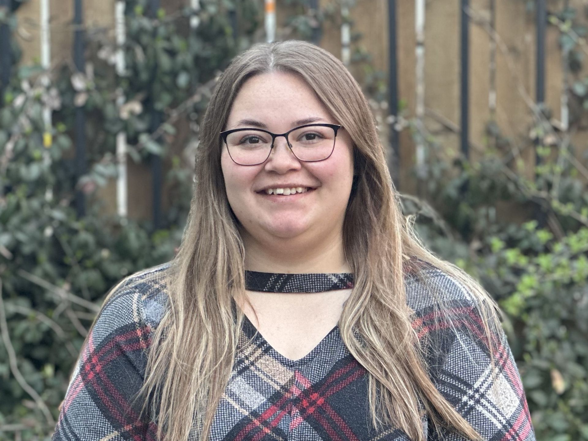 Smiling person with glasses in a plaid top, standing outdoors in front of greenery and a wooden fence.