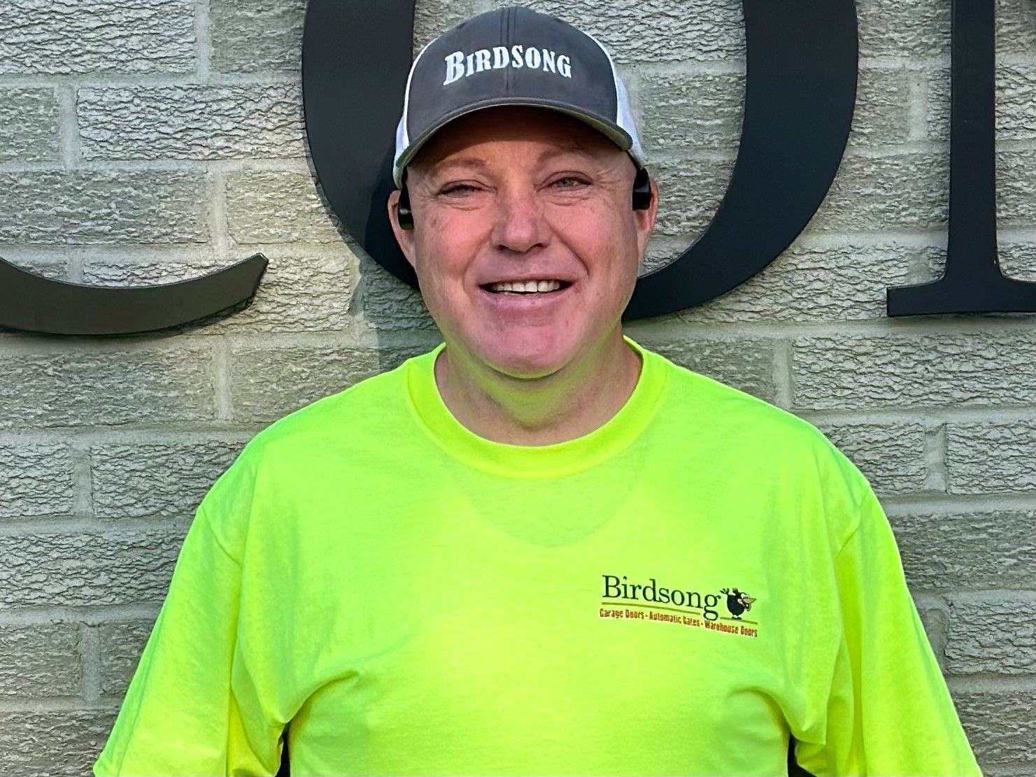 Smiling person in a neon yellow shirt and black cap standing before a stone wall