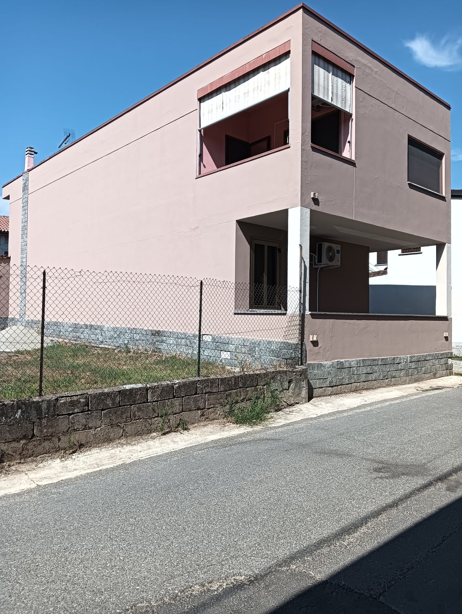Casa rosa a due piani in una strada tranquilla con un basso muro di pietra e cielo azzurro.