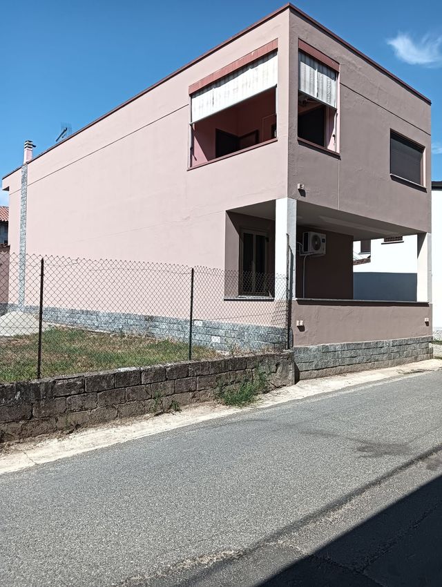 Casa rosa a due piani in una strada tranquilla con un basso muro di pietra e cielo azzurro.
