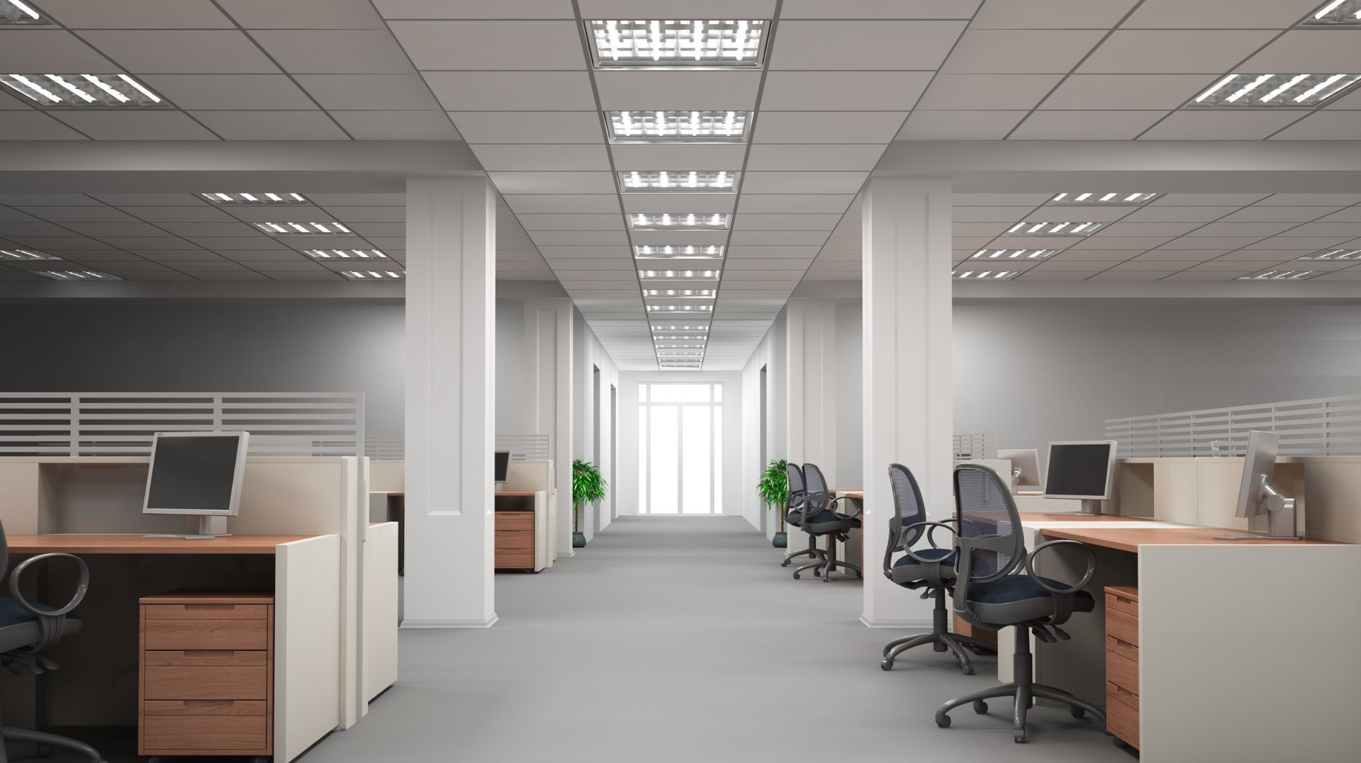Empty modern office with rows of desks, computers, and gray carpet under bright fluorescent ceiling lights.