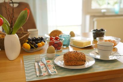 Frühstückstisch mit Kaffee, Gebäck, Obst und einer Blumenvase an einem sonnendurchfluteten Fenster