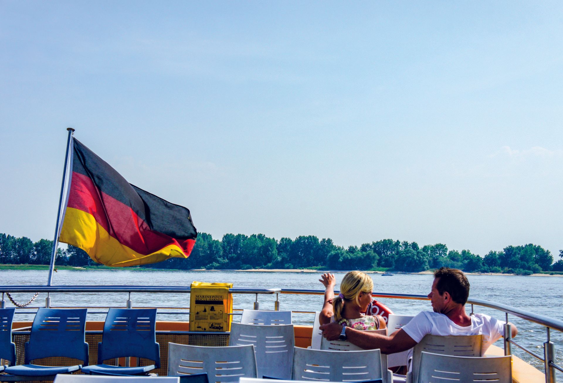 Oberdeck einer Fähre mit Deutschlandfahne, blauen Stühlen und zwei Personen, die am Wasser sitzen