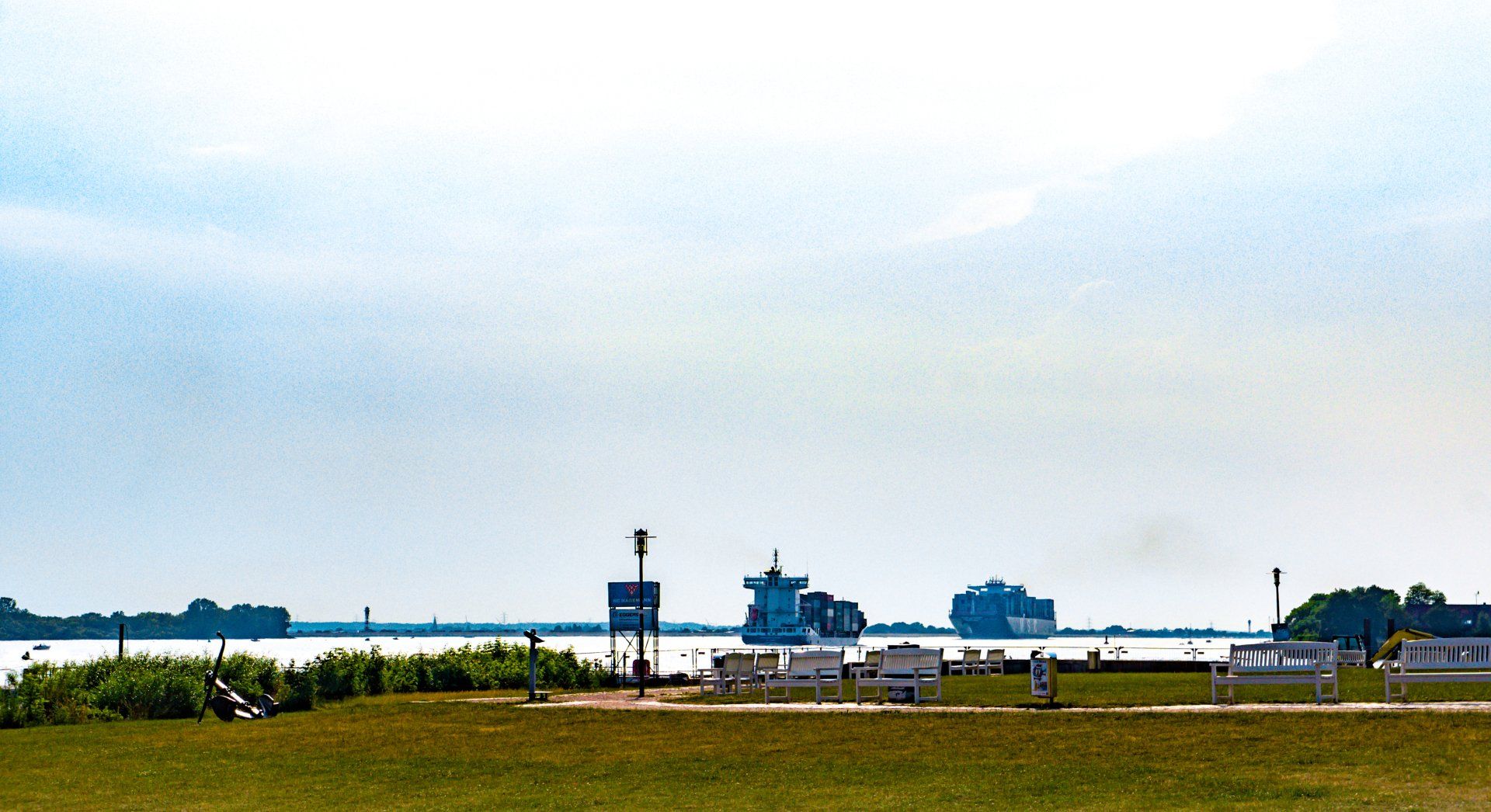 Strandpromenade mit grasbewachsenem Feld und Schiffen auf der Elbe unter einem hellen, bewölkten Himmel