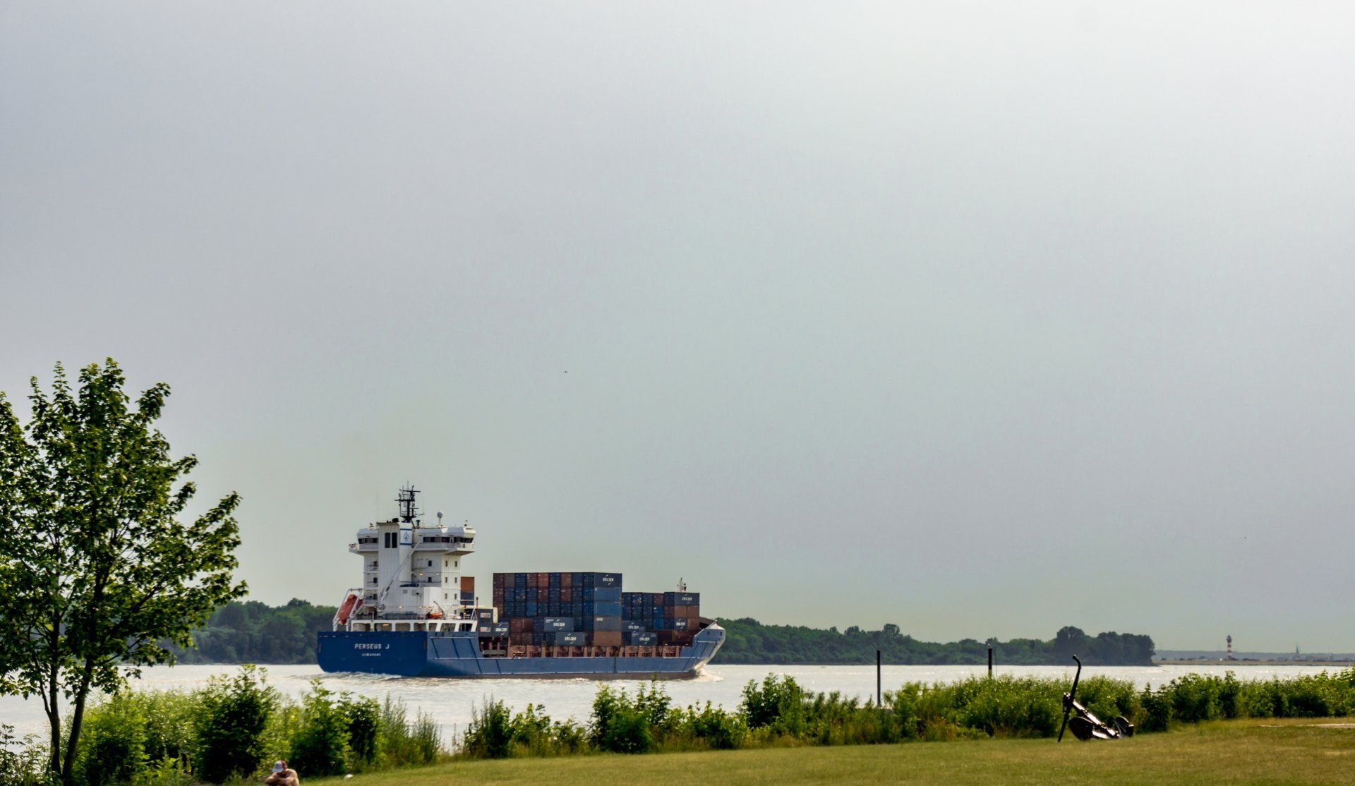 Frachtschiff auf ruhigem Wasser nahe einer grasbewachsenen Küste unter bewölktem Himmel