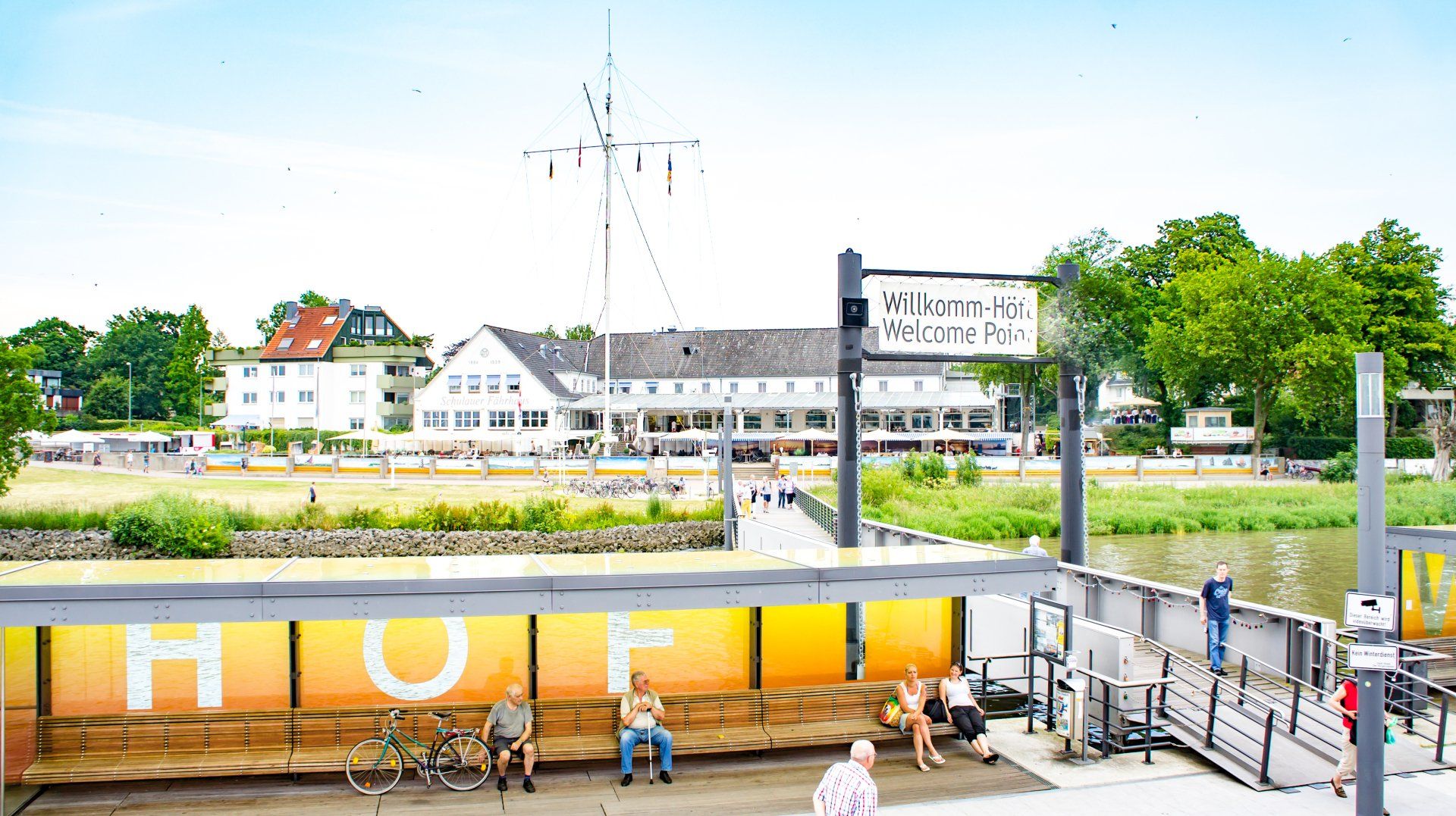 Café am Elbufer mit orangefarbenen Sitzgelegenheiten, Menschen und einem weißen Gebäude auf der anderen Wasserseite