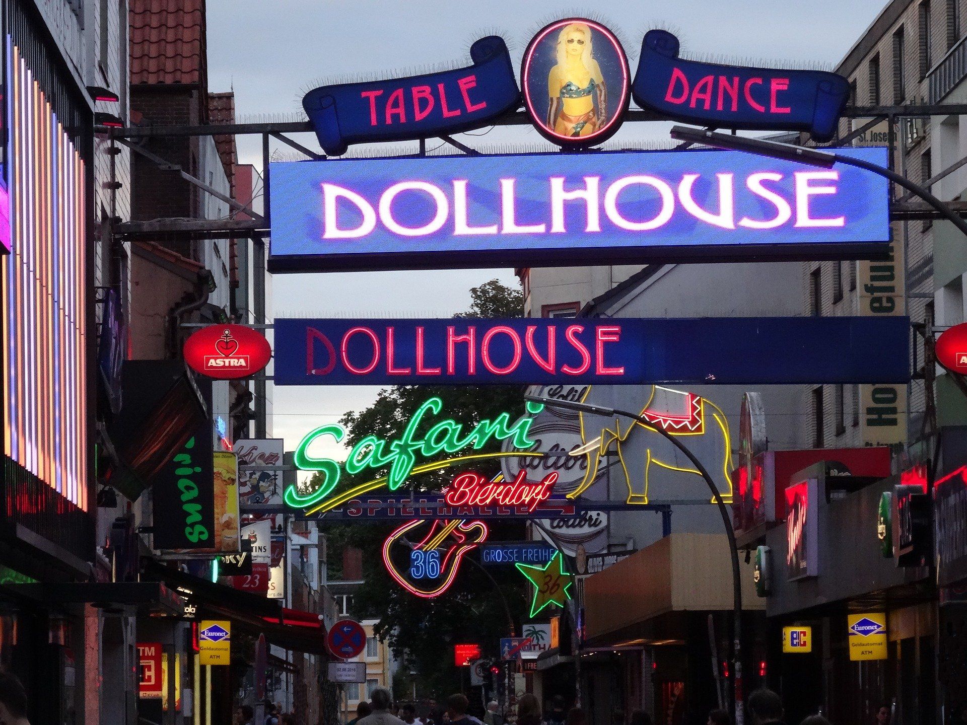 Eine neonbeleuchtete Straße mit einem leuchtenden „DOLLHOUSE“-Schild über einer belebten Gasse mit Nachtleben.